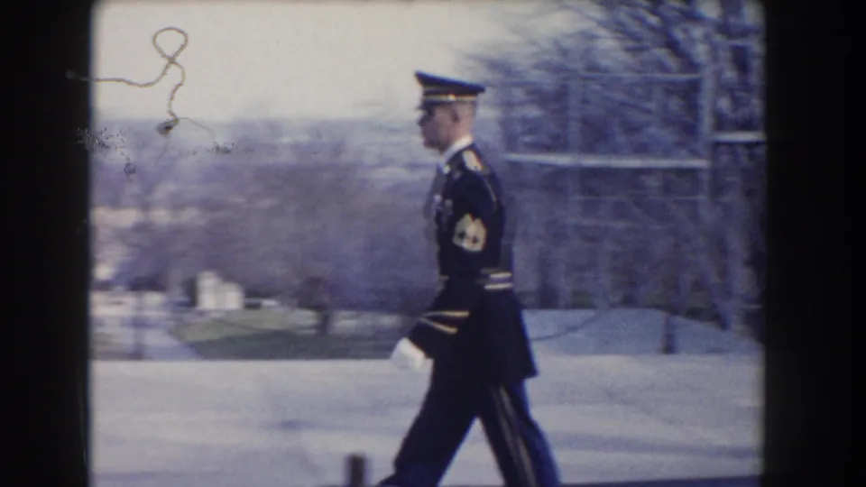 A uniformed guard in white gloves and peaked cap marches in profile near a pale stone monument structure in Washington, DC.