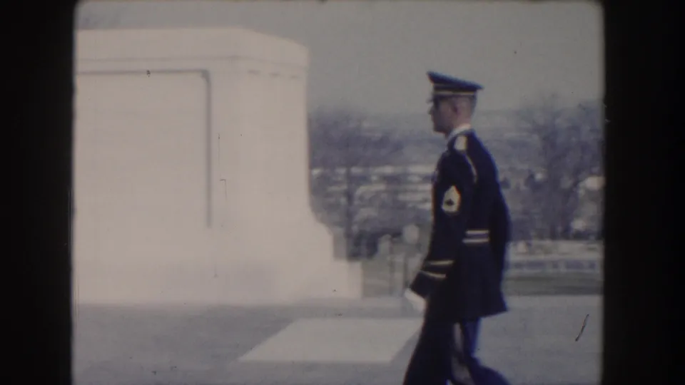 A guard in dark ceremonial uniform walks past a pale monument wall, with Washington, DC scenery faintly visible beyond.