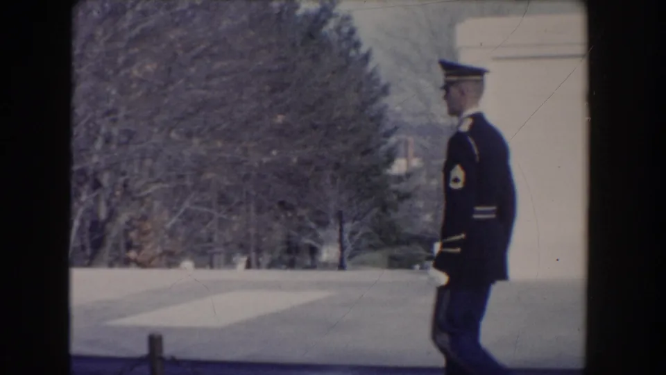 A uniformed guard appears near the edge of a pale stone monument structure, with bare trees across the plaza in Washington, DC.