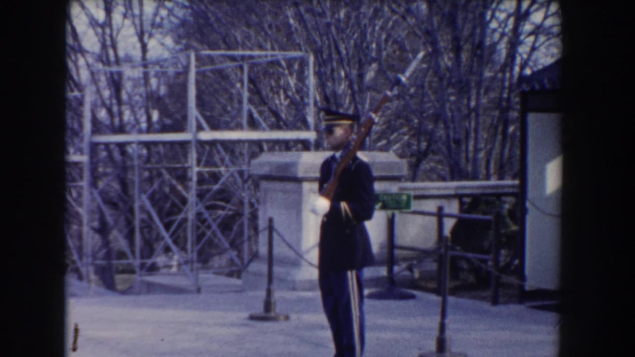 Uniformed soldier holding a rifle near a stone memorial area and metal scaffolding.