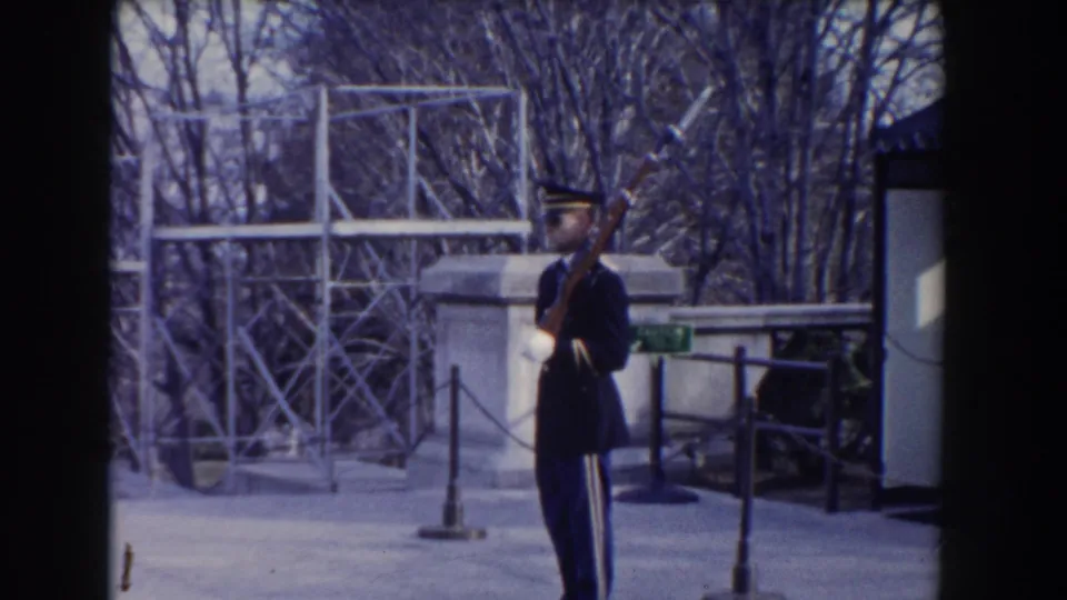 A uniformed soldier in a dark dress uniform holds a rifle near the Tomb of the Unknown Soldier setting in the supplied 1960 Washington, DC footage.