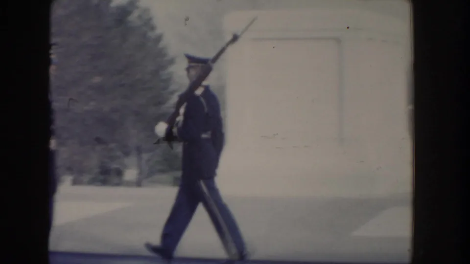 A soldier in dark ceremonial uniform marches with a rifle near a pale stone memorial wall in the supplied 1960 Washington, DC footage.