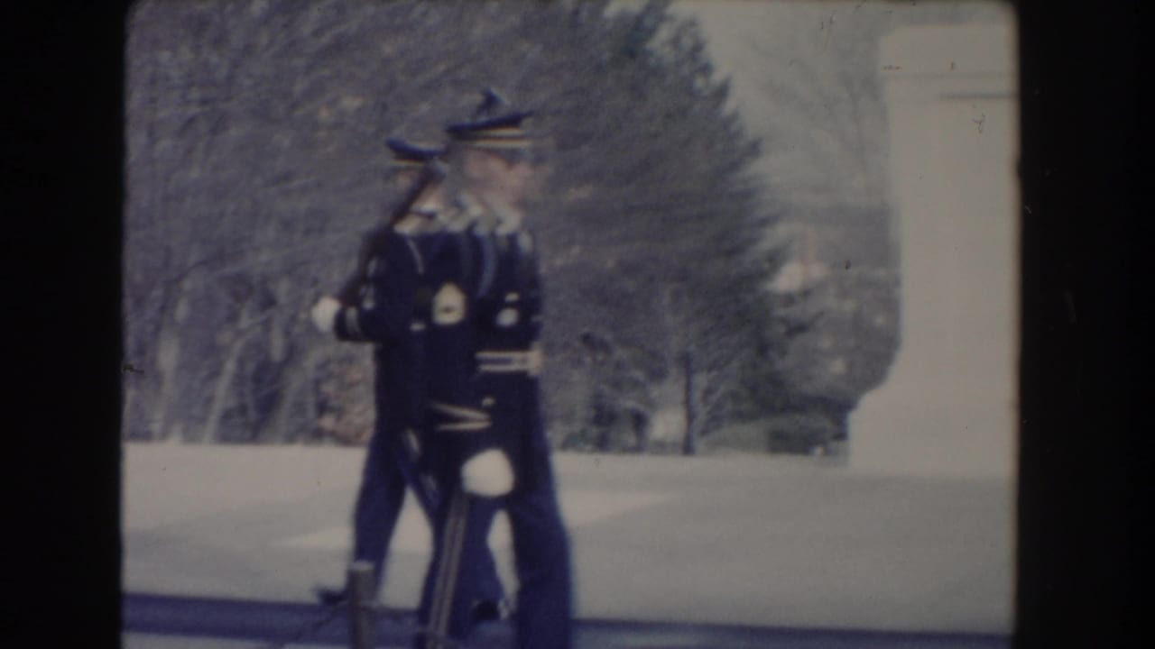 Uniformed guards with rifles march beside a stone monument base.