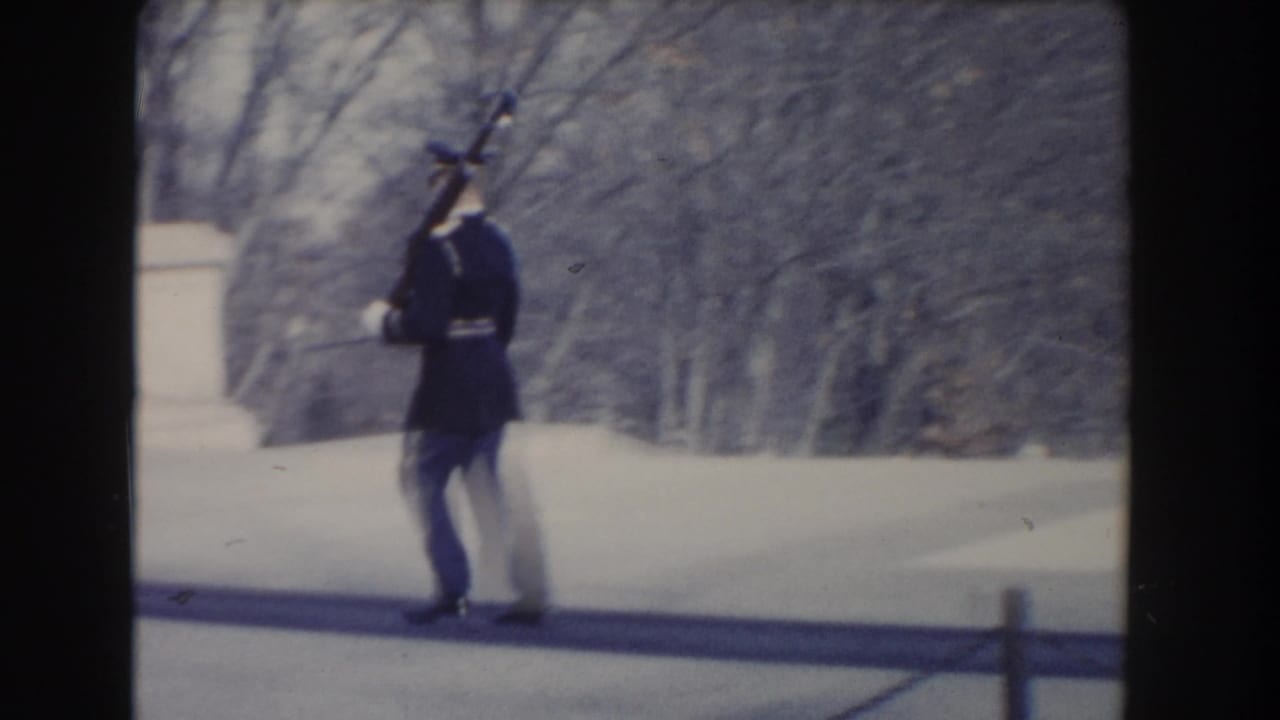 A uniformed guard in the Washington, DC clip walks with a rifle near stonework and winter-bare trees.