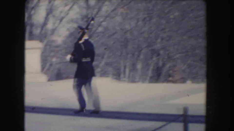 A uniformed guard in the Washington, DC clip walks with a rifle near stonework and winter-bare trees.