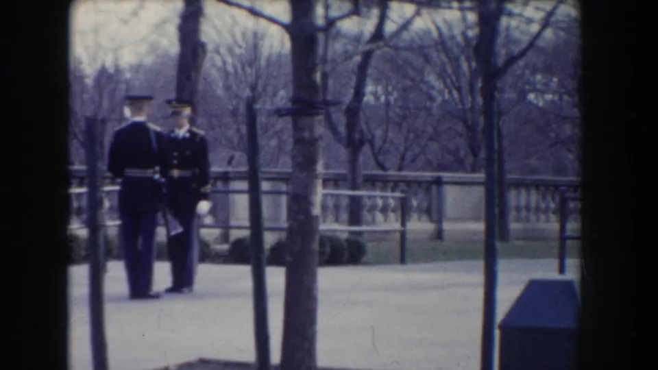 Two uniformed guards appear to speak or exchange positions beside trees and a stone railing in Washington, DC.