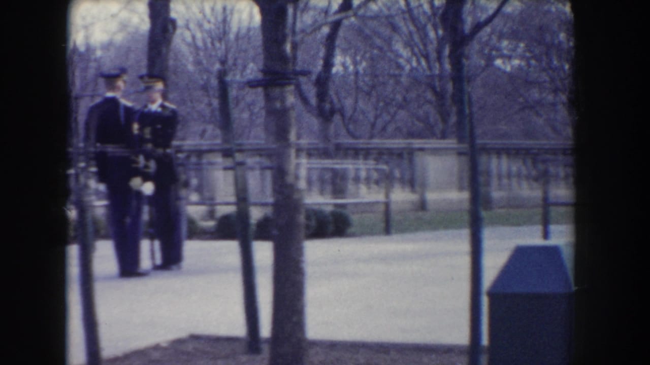 In Washington, DC circa 1960, two dress-uniformed military figures stand near a stone balustrade in a leafless park setting.