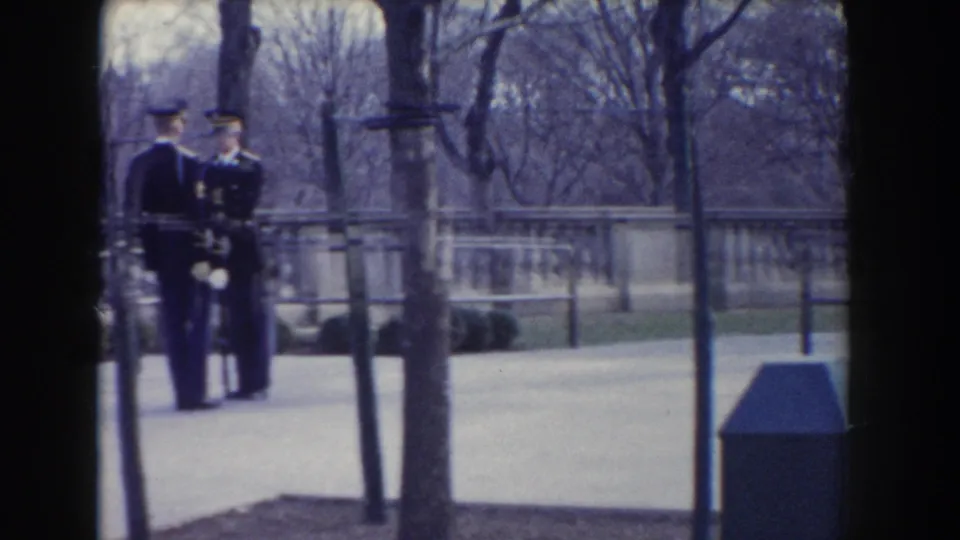 In Washington, DC circa 1960, two dress-uniformed military figures stand near a stone balustrade in a leafless park setting.