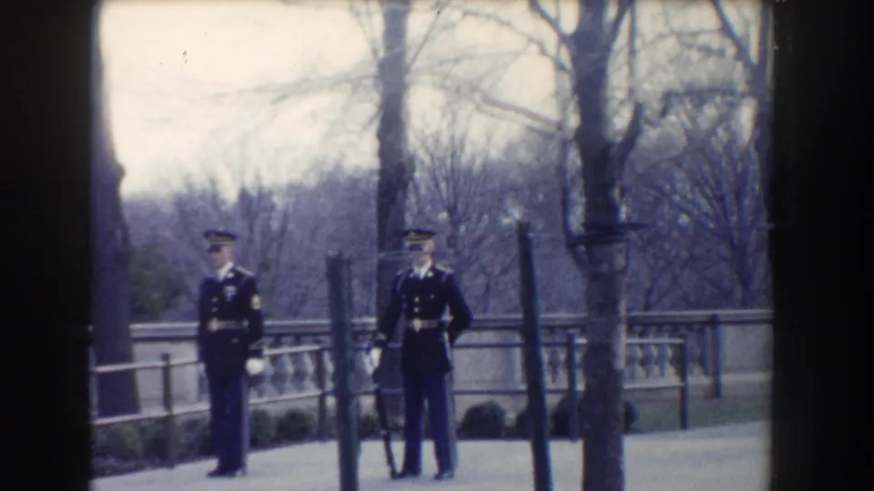 In a Washington, DC park-like setting, two uniformed guards stand separated near a tree and stone balustrade.