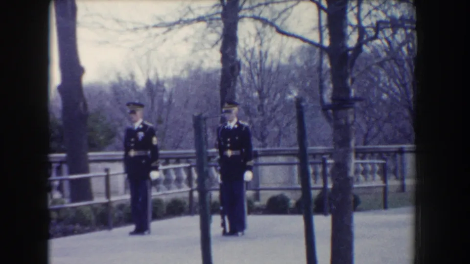 Two formal military guards stand on a Washington, DC walkway with bare trees and a stone railing behind them.