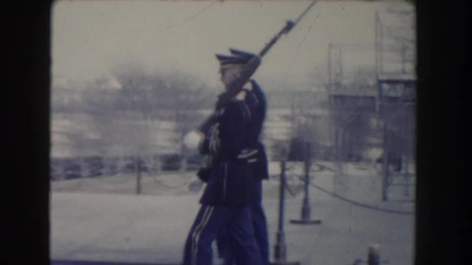 A Washington, DC honor guard in dark uniform and white gloves marches with a rifle and fixed bayonet beside snow and winter trees.