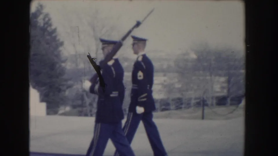 Two honor guards in formal uniforms pass in formation in Washington, DC, with one carrying a rifle and bayonet across a snowy paved area.