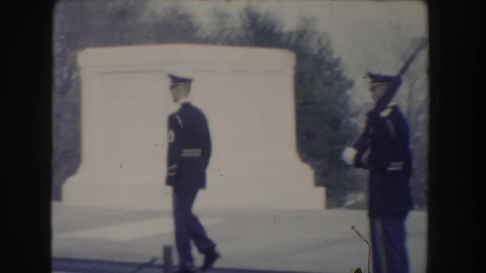 One honor guard walks before a white stone monument while another uniformed guard with a rifle stands at the edge of the frame.