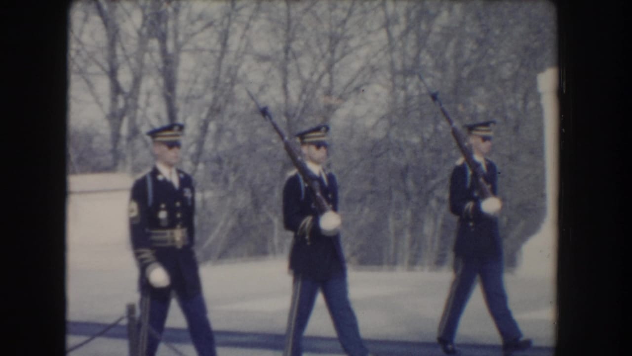 The honor guard formation advances past the marble memorial with rifles raised and bare trees behind them.