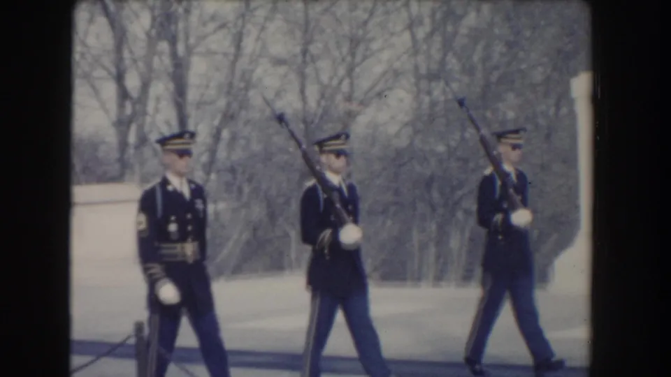 The honor guard formation advances past the marble memorial with rifles raised and bare trees behind them.