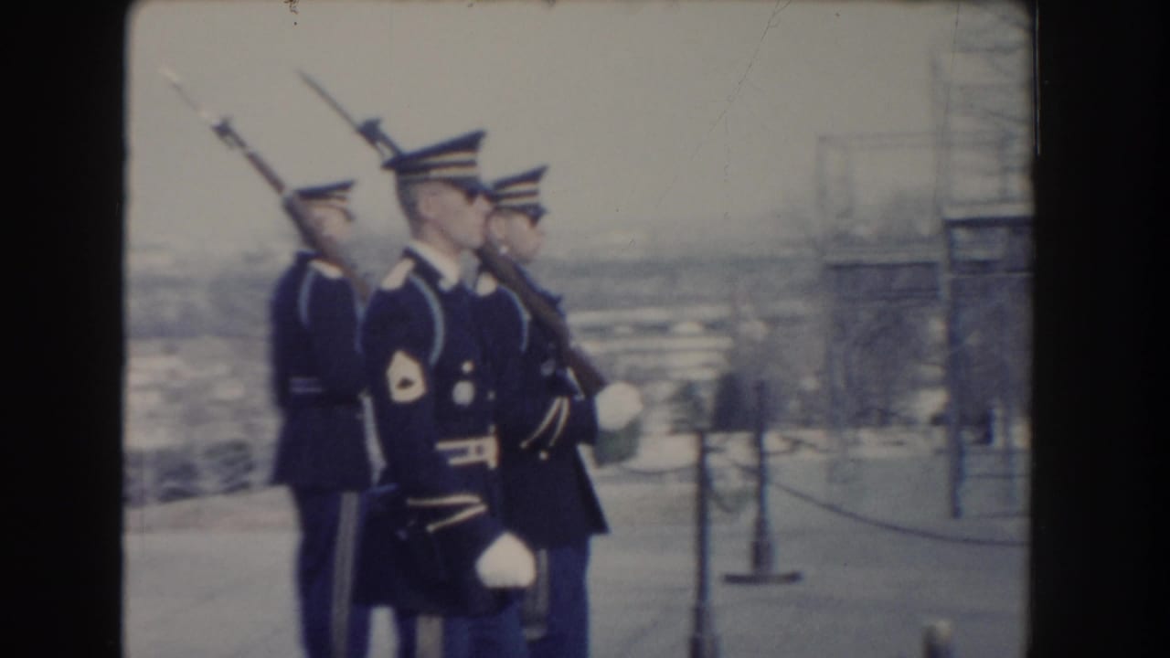 Uniformed soldiers with rifles and bayonets march near a stone wall and chain barrier.