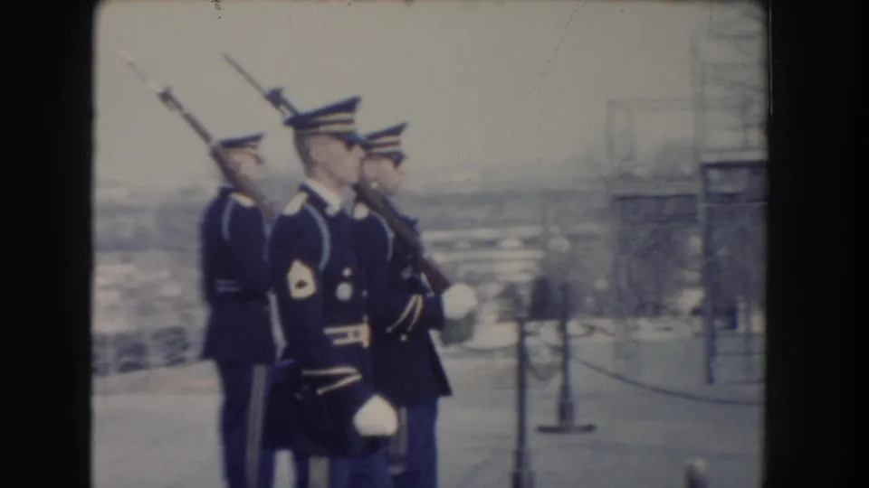 In 1960 Washington DC footage, uniformed soldiers carry rifles with bayonets while marching past a stone wall and visitor barriers.