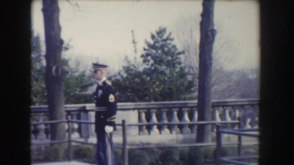 The home-movie frame holds on a uniformed soldier at attention beside a tree-lined stone terrace in Washington DC, 1960.