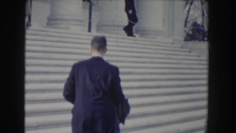 A man in a dark suit climbs broad stone steps from behind in 1960 Washington DC footage, with another figure near the upper landing.