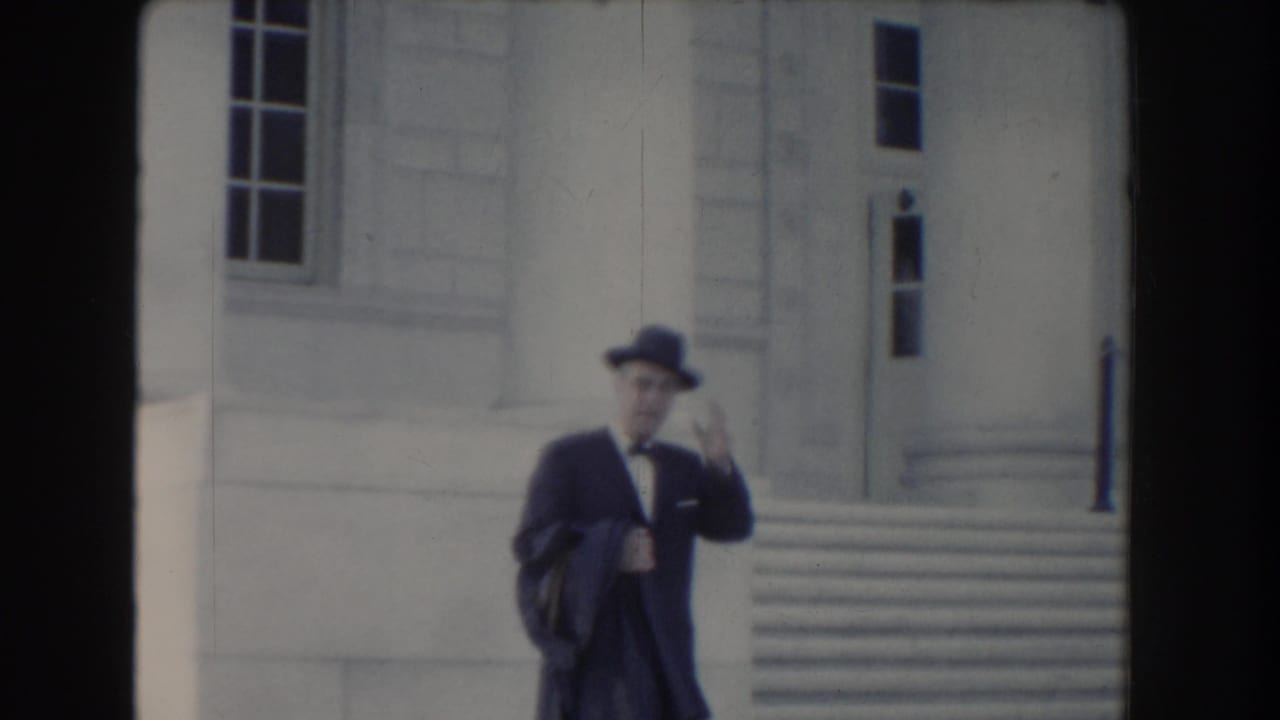 A suited man with a brimmed hat and glasses passes the stone facade of a Washington DC civic building in 1960 footage.