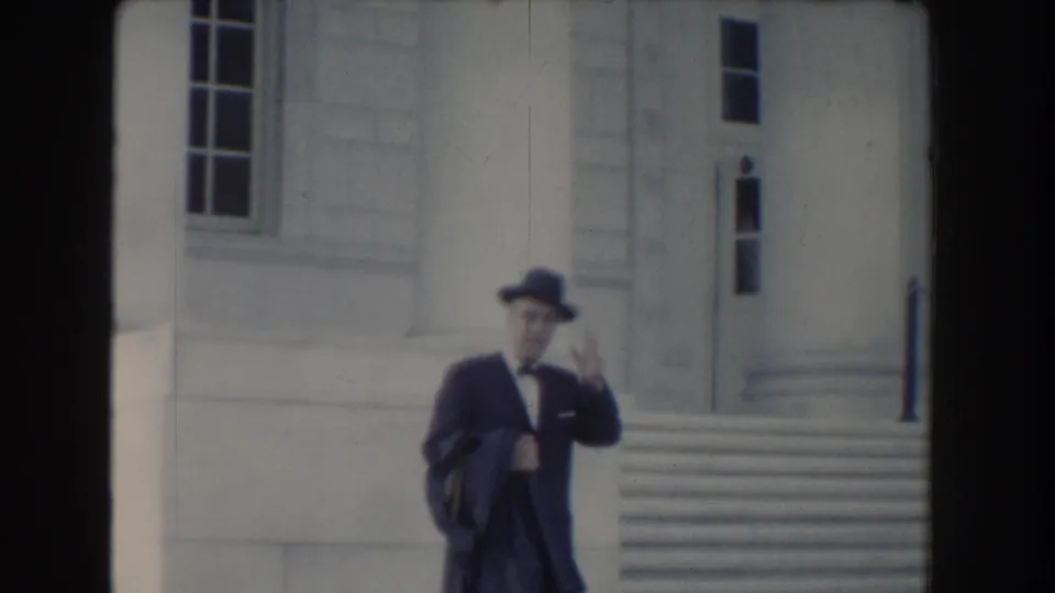 A suited man with a brimmed hat and glasses passes the stone facade of a Washington DC civic building in 1960 footage.