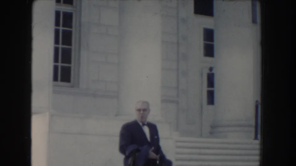 A suited man with glasses is framed against a pale stone government-style exterior in Washington DC archival 8mm footage.