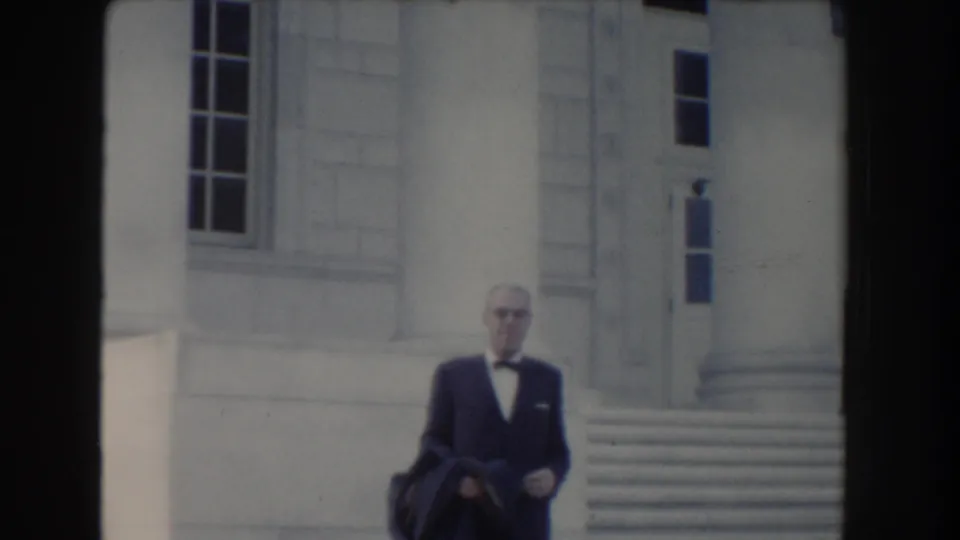 A man in a dark suit and glasses faces the camera area near civic-building steps in 1960 Washington DC footage.