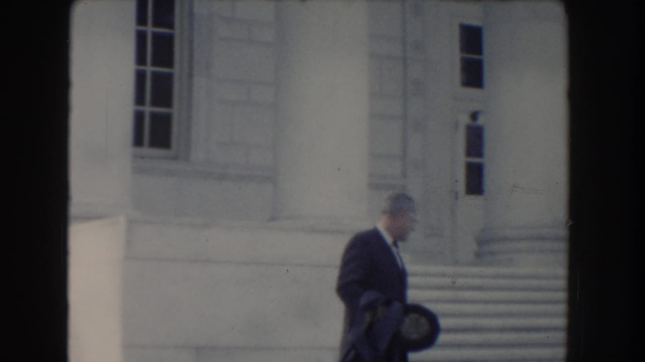 The man in formal dress moves right across the frame outside a columned building in Washington, DC home-movie footage.