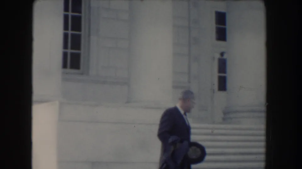 The man in formal dress moves right across the frame outside a columned building in Washington, DC home-movie footage.