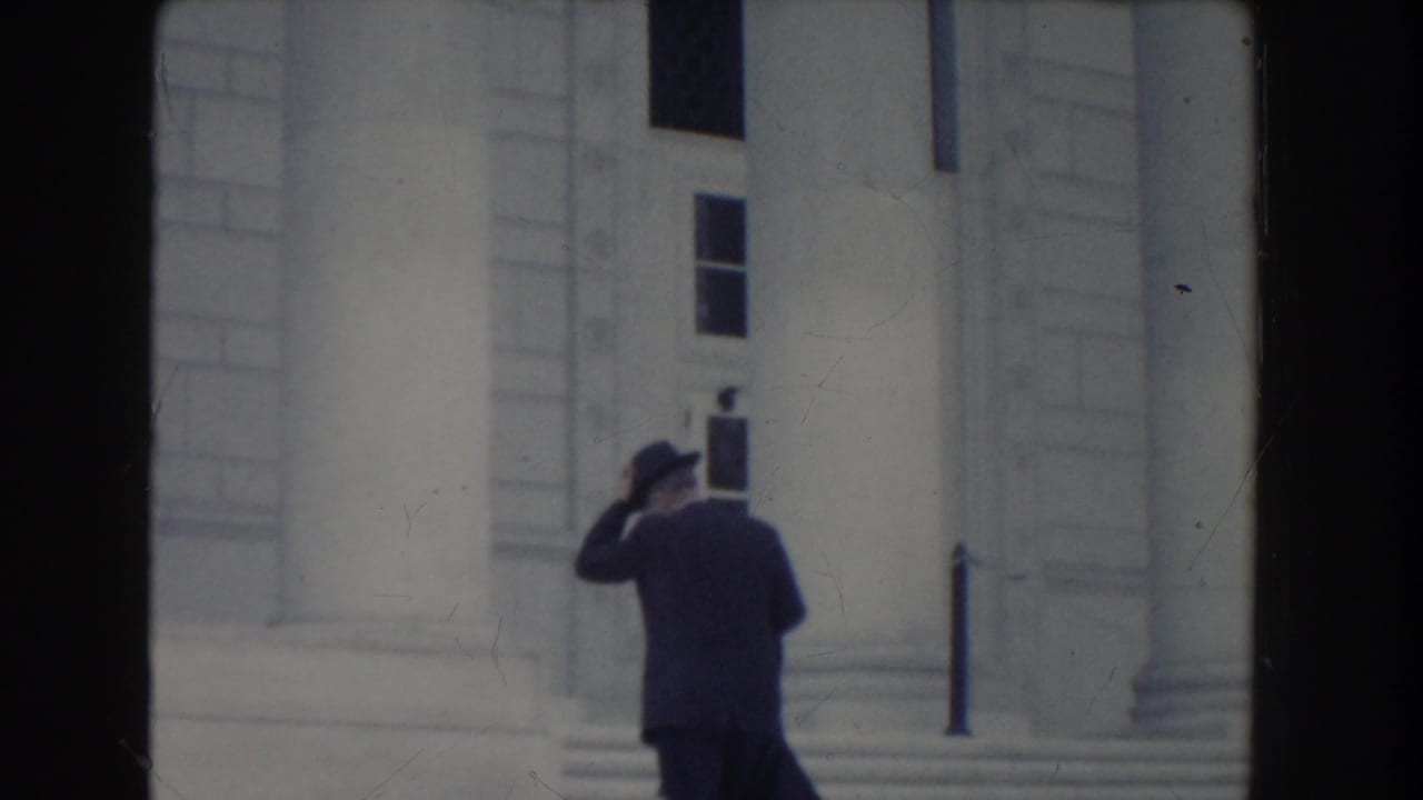 In Washington DC 1960 home-movie footage, a suited man in a fedora adjusts his hat while walking toward a white columned building.