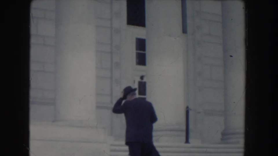 In Washington DC 1960 home-movie footage, a suited man in a fedora adjusts his hat while walking toward a white columned building.