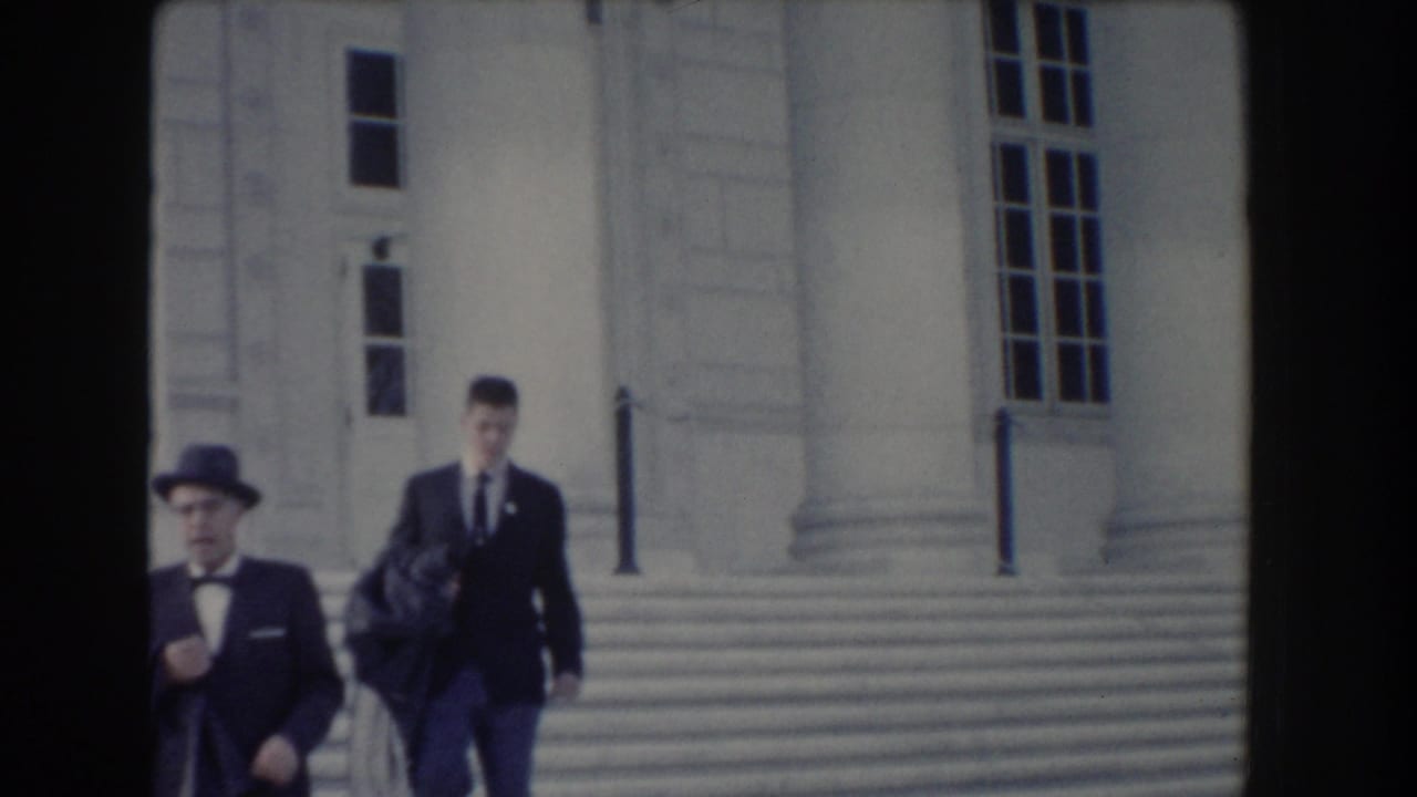 Two men in dark suits walk down broad stone steps outside a columned building.