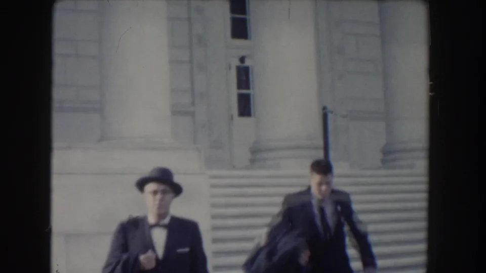 Two men in formal clothing move down stone steps in Washington, DC, one wearing a brimmed hat and bow tie.