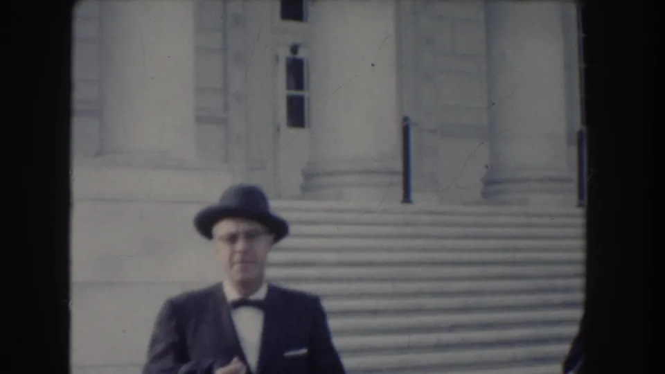 A formally dressed man with glasses, bow tie, and brimmed hat approaches the camera near broad stone steps in Washington, DC.