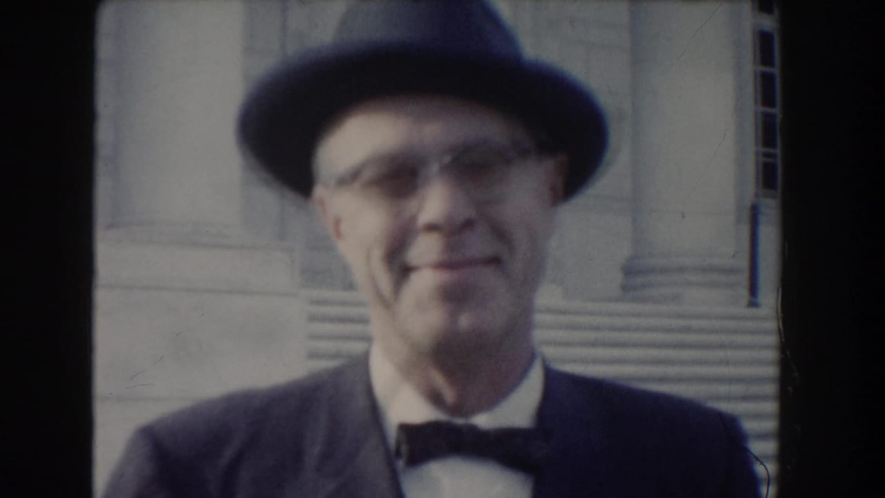 In 1960 Washington, DC footage, an adult man in a dark fedora, suit, and bow tie smiles toward the camera near stone steps.