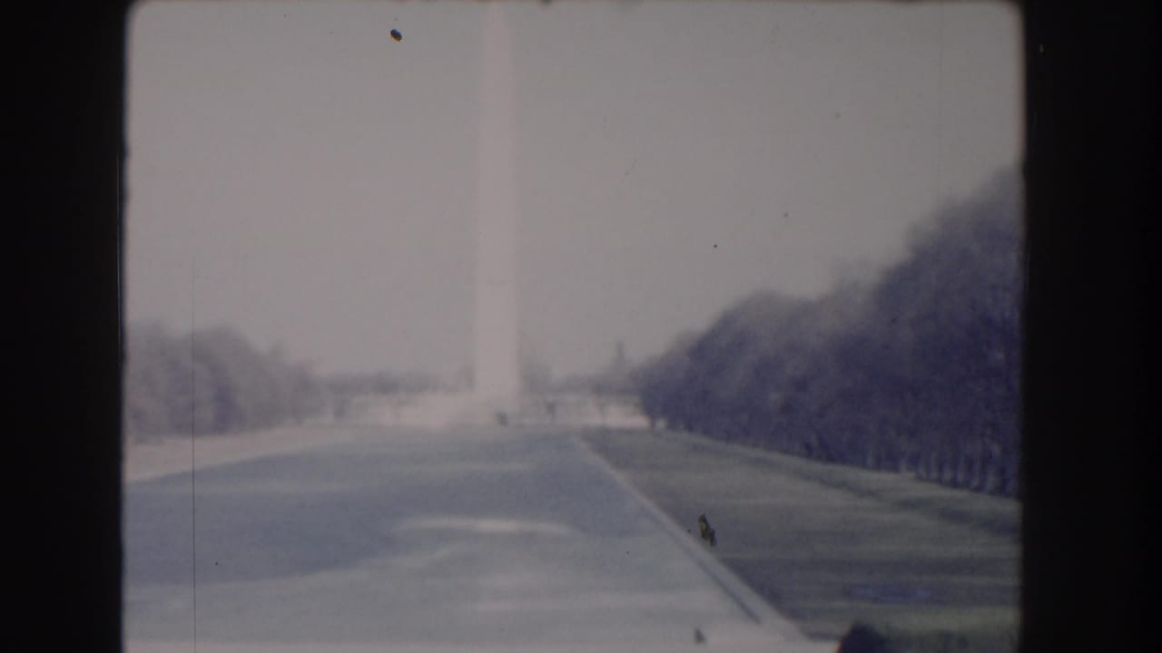 1960 Washington, DC home-movie frame showing the Washington Monument in haze beyond a broad road and tree-lined lawns.