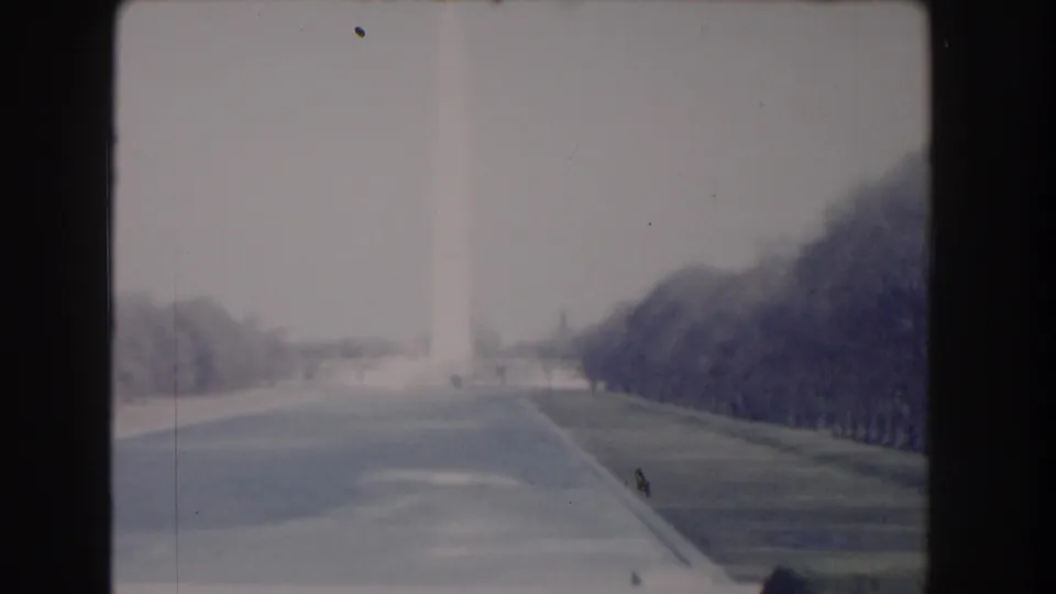 1960 Washington, DC home-movie frame showing the Washington Monument in haze beyond a broad road and tree-lined lawns.