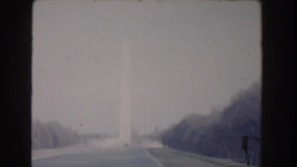 Hazy 1960 Washington, DC home-movie view of the monument from a road flanked by lawns and trees.