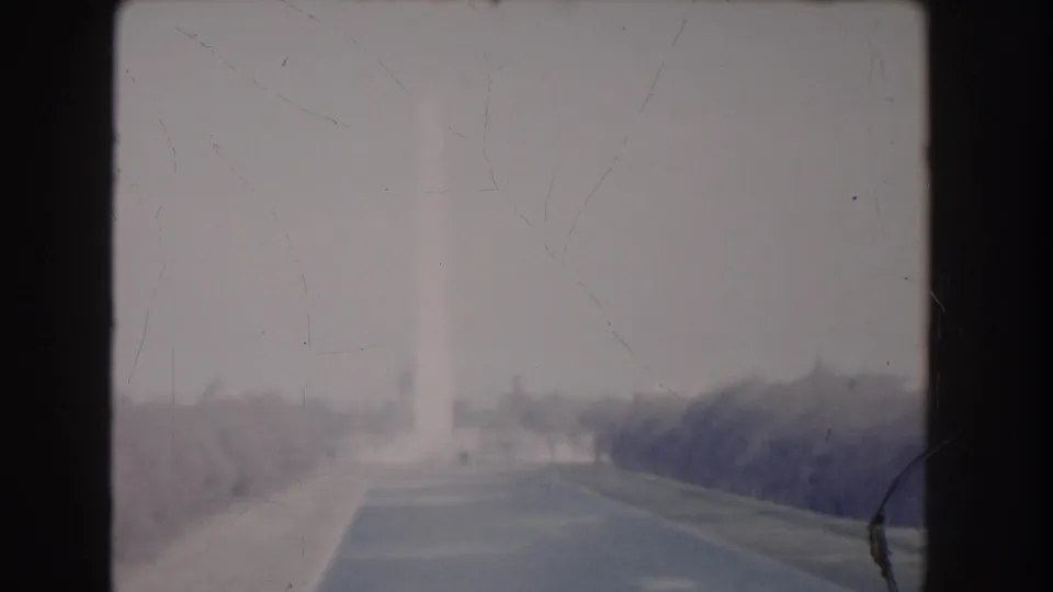 1960 Washington DC frame with the Washington Monument faintly visible beyond a tree-lined road under overexposed light.