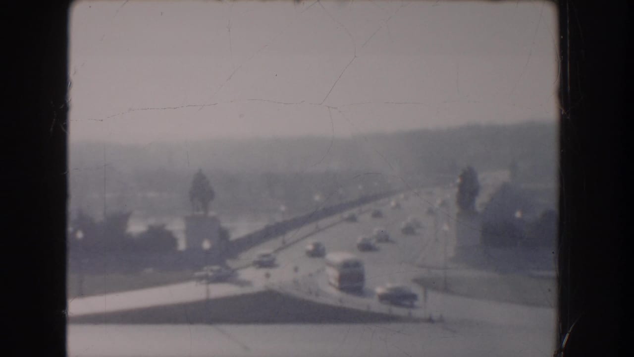 1960 Washington DC home-movie frame showing light traffic on a bridge or elevated roadway, with water, trees, and haze in the distance.