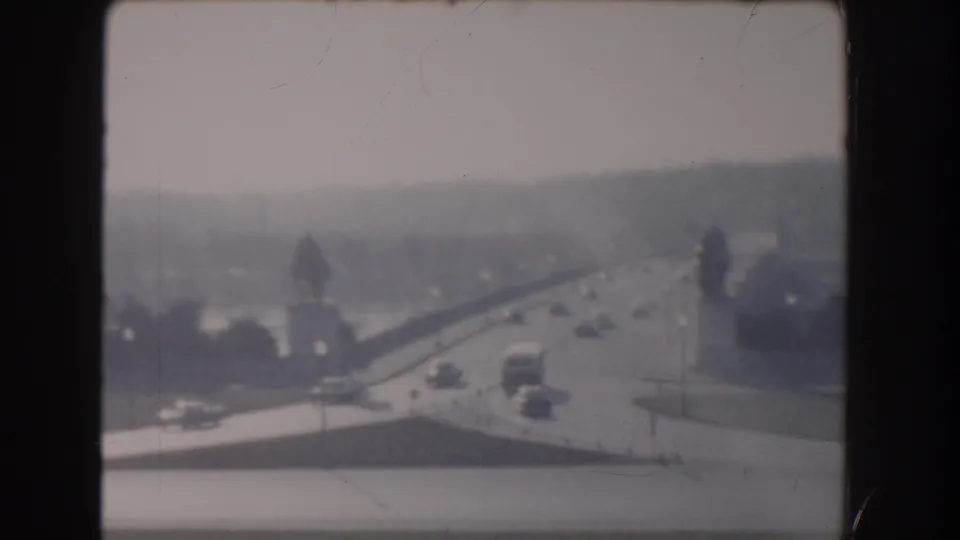 High-angle 1960 Washington DC view of cars moving along a bridge or roadway near water, with a distant tree line under soft daylight.