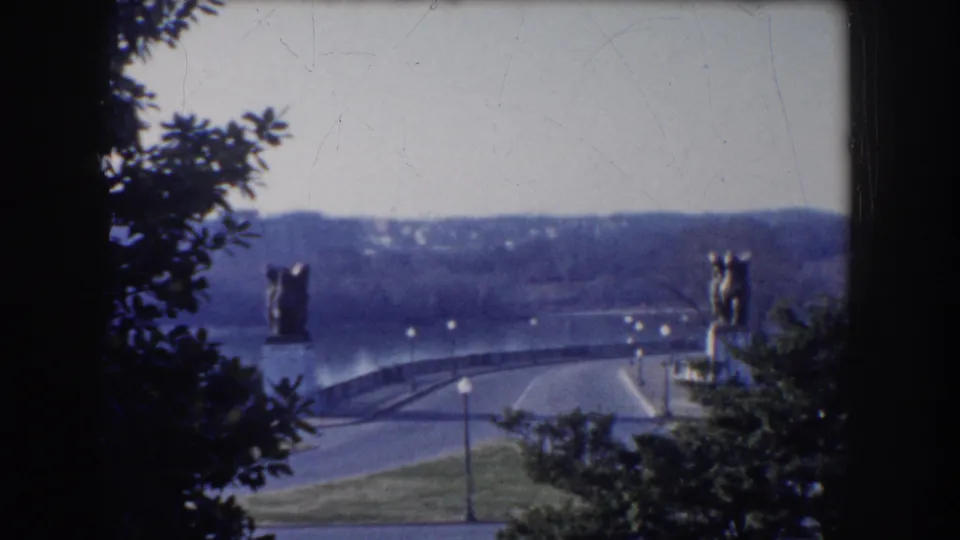 Washington DC overlook with green grass, water, a road bridge, trees, and distant sculptural figures in a 1960 8mm home-movie frame.