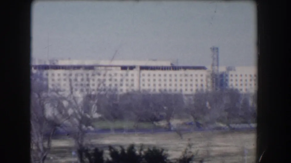 1960 Washington DC frame with a tan multi-story building across open grass, bare trees, and a pale sky, filmed from a steady elevated viewpoint.