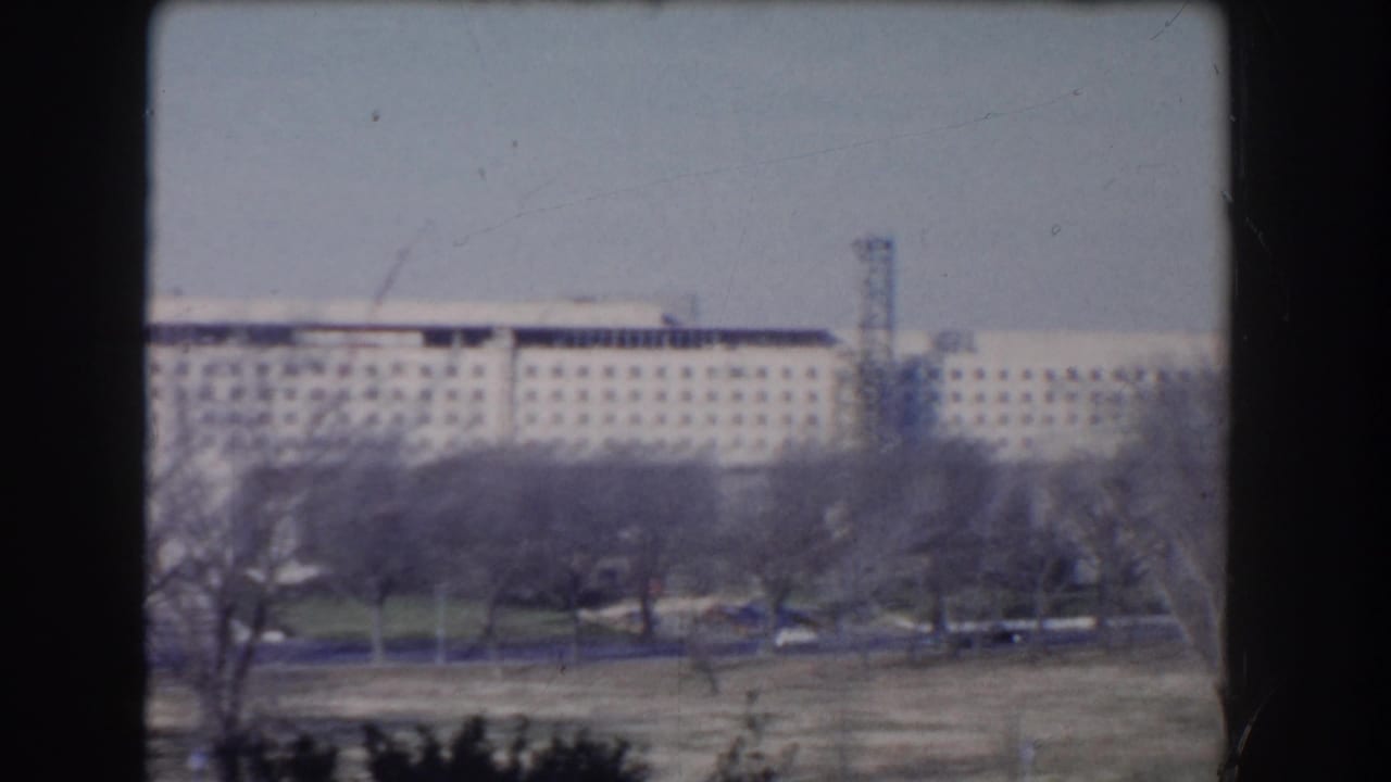 Distant institutional building across trees and open ground, seen through 8mm frame edges.