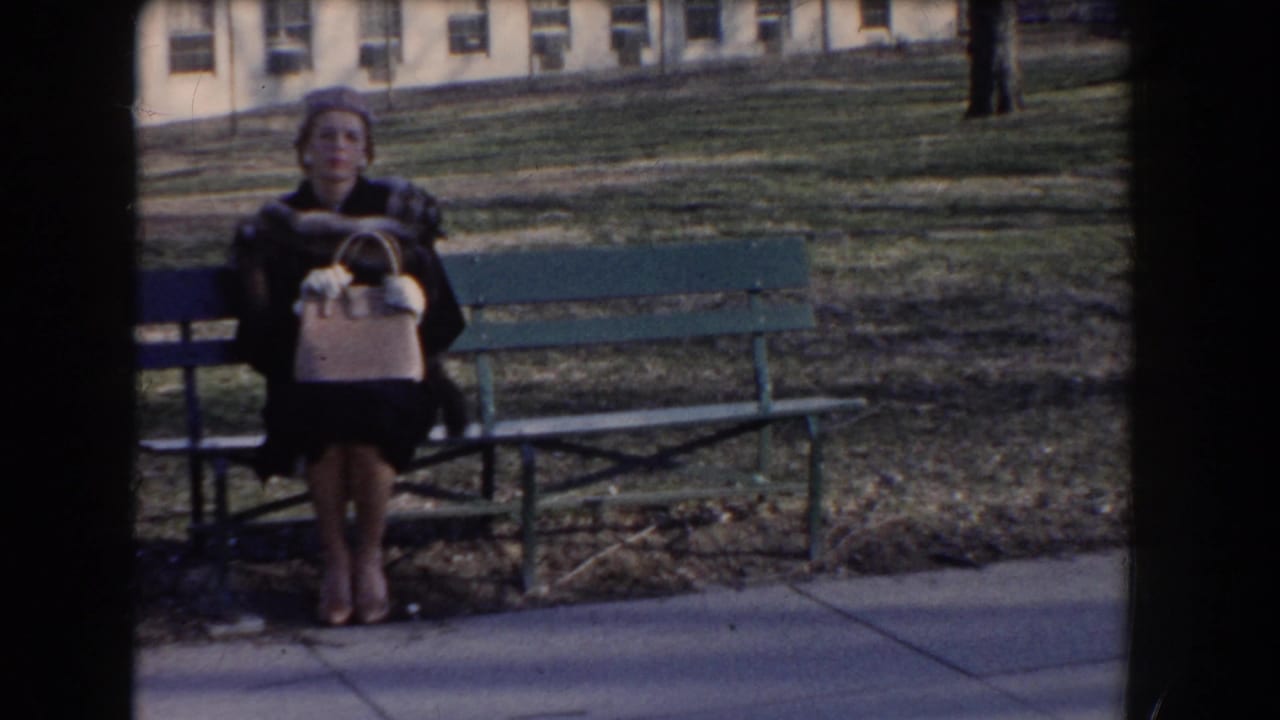 Woman in dark coat and hat seated on a green bench holding a light purse.