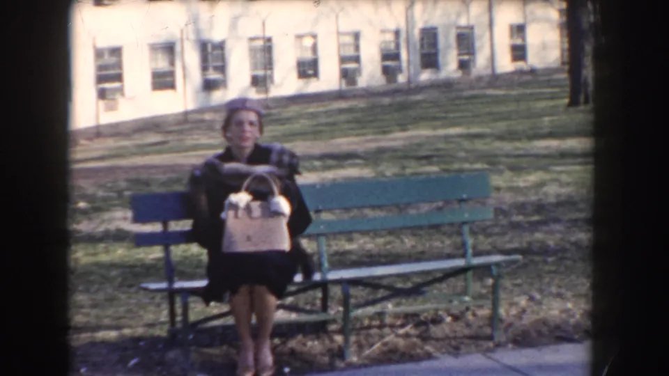 Home-movie frame showing the same seated adult woman on a park bench, with lawn and building windows in the background.