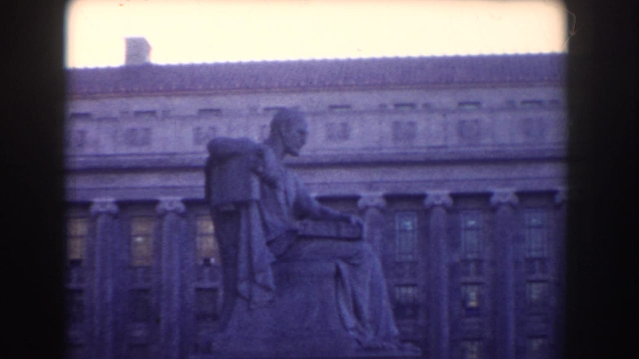 Side view of a seated statue before a large columned building.