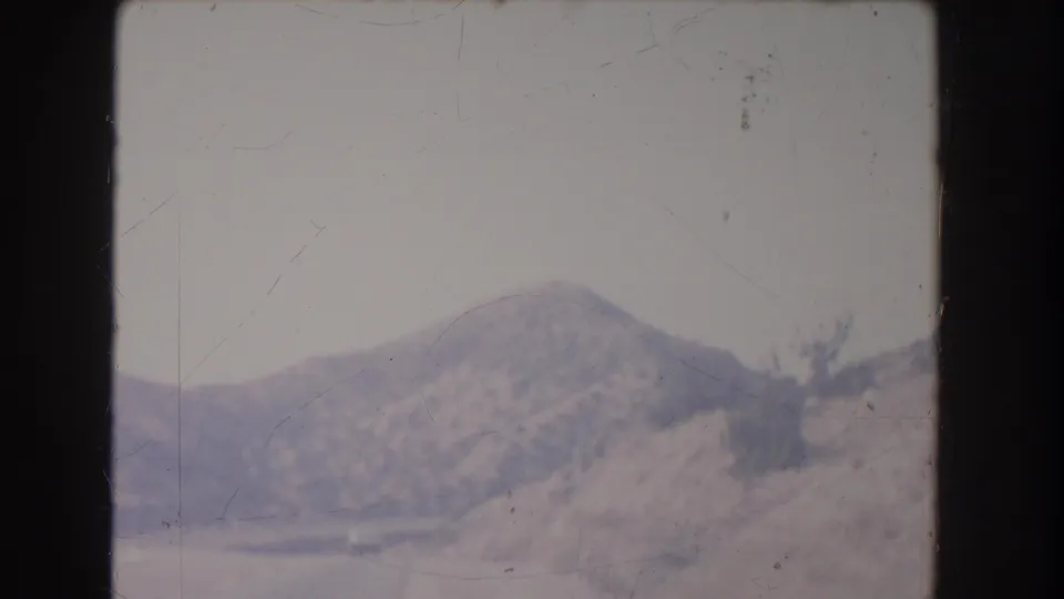 Soft 8mm roadside travel frame showing dry rolling hills, sparse vegetation, and a pale sky from a moving-vehicle perspective.
