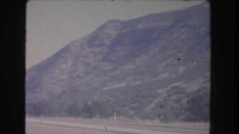 Faded 8mm view of a mountain slope beside a road, with sparse trees and dry terrain visible through soft focus.
