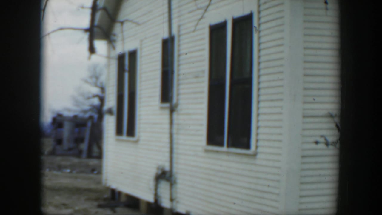 1951 Cuba home-movie frame showing a white clapboard house exterior with dark windows, utility pipe, and bare branches.
